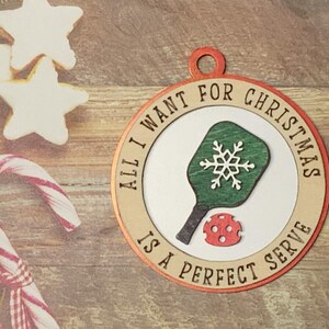 May include: A round wooden ornament with a red and white border. The ornament features a green and white pickleball paddle with a snowflake design and the text "All I want for Christmas is a perfect serve".