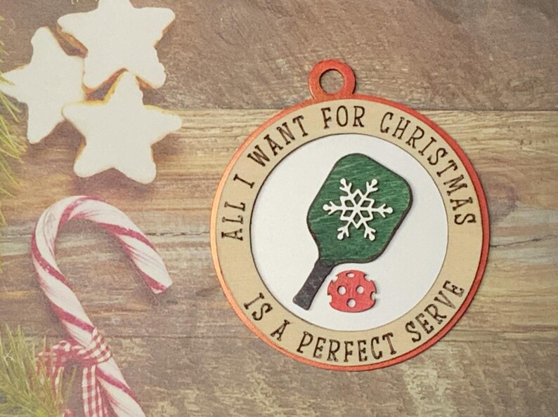 May include: A round wooden ornament with a red and white border. The ornament features a green and white pickleball paddle with a snowflake design and the text "All I want for Christmas is a perfect serve".