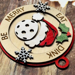 May include: A wooden Christmas ornament with a red, white, and green design. It features a Santa hat, a snowflake, and a pickleball. The ornament reads "Be Merry Eat Dink".