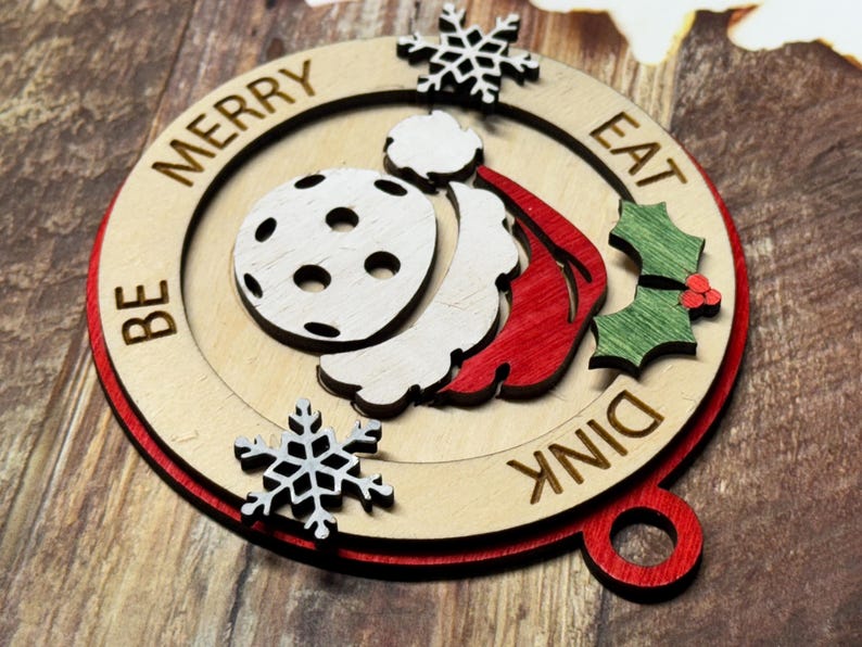 May include: A wooden Christmas ornament with a red, white, and green design. It features a Santa hat, a snowflake, and a pickleball. The ornament reads "Be Merry Eat Dink".
