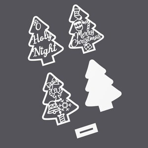Op de afbeelding: Vier witte kerstboomuitsparingen met verschillende ontwerpen. Eén boom zegt "O Holy Night", een andere zegt "Merry Christmas", één heeft een sneeuwvlok, bel, engel en kaars, en de laatste is leeg.