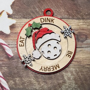 May include: A wooden Christmas ornament shaped like a circle with a red border. The ornament features a white and gray pickleball with a red Santa hat on top. The text "DINK EAT BE MERRY" is written around the pickleball.