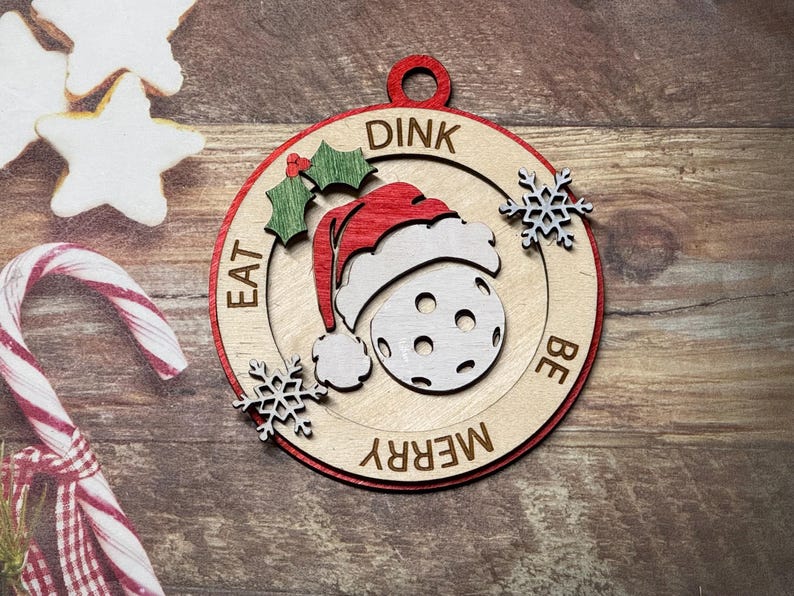 May include: A wooden Christmas ornament shaped like a circle with a red border. The ornament features a white and gray pickleball with a red Santa hat on top. The text "DINK EAT BE MERRY" is written around the pickleball.