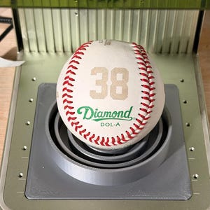 May include: A baseball with red stitching and the number 38 printed in beige. The word "Diamond" is printed in green, and the ball is resting on a gray, circular platform.