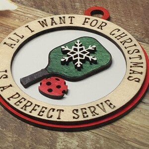 May include: A wooden Christmas ornament with a red border. It features a green and white pickleball paddle with a snowflake design and the text "All I want for Christmas is a perfect serve".