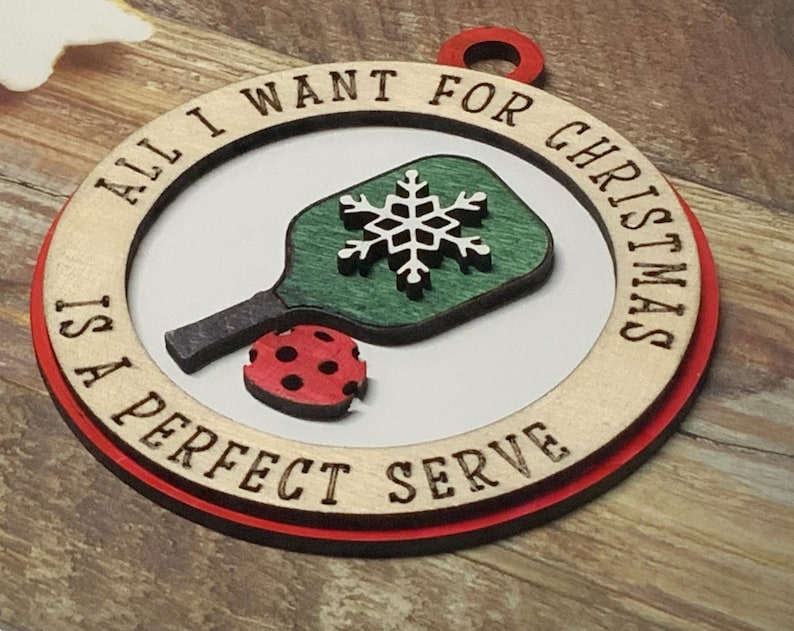 May include: A wooden Christmas ornament with a red border. It features a green and white pickleball paddle with a snowflake design and the text "All I want for Christmas is a perfect serve".