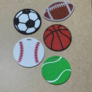 May include: Five round wooden ornaments with different sports ball designs. The ornaments are white with black, brown, red, and green designs. The sports balls include a soccer ball, football, basketball, baseball, and tennis ball.