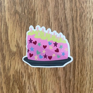 May include: A pink cake with green frosting and colorful sprinkles in the shape of stars and hearts. The cake is on a black plate.