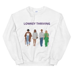 以下が含まれることがあります： 紫色の文字で「Lowkey Thriving」と書かれた白いスウェットシャツ。スウェットシャツには、一緒に歩いている4人の女性のイラストが描かれています。