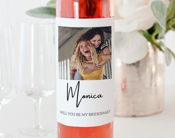 Op de afbeelding: Een roze wijnfles met een wit etiket. Het etiket heeft een foto van twee vrouwen die lachen en de tekst "Monica Wil je mijn bruidsmeisje zijn?"