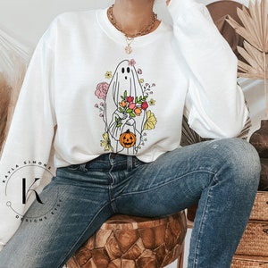 May include: White sweatshirt with a graphic of a ghost holding a pumpkin and flowers. The ghost is outlined in black and the flowers are in shades of pink, yellow, and orange.