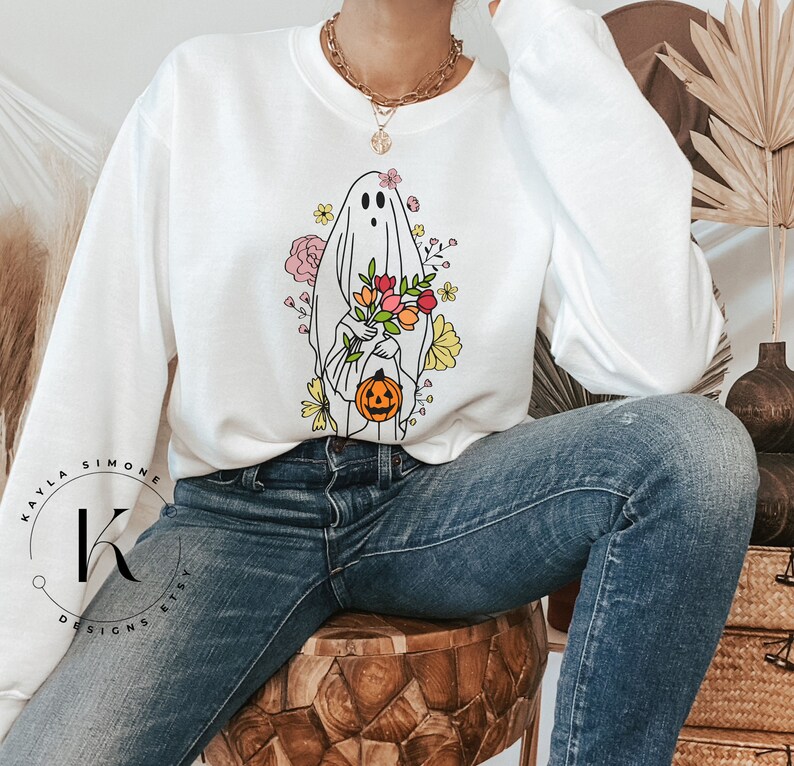 May include: White sweatshirt with a graphic of a ghost holding a pumpkin and flowers. The ghost is outlined in black and the flowers are in shades of pink, yellow, and orange.