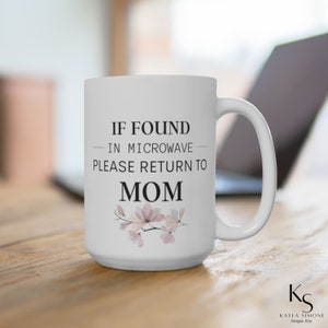 May include: White ceramic mug with a black and white design that says "If found in microwave please return to Mom". The mug has a floral design with pink and white flowers.