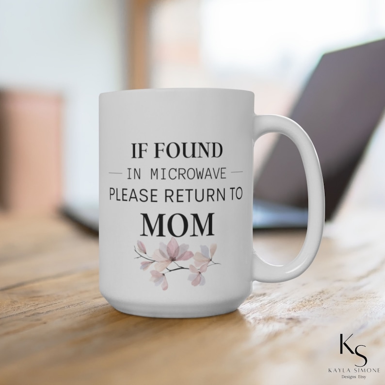 May include: White ceramic mug with a black and white design that says "If found in microwave please return to Mom". The mug has a floral design with pink and white flowers.