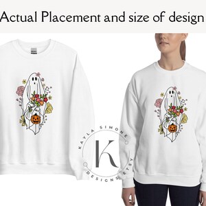 May include: White sweatshirt with a graphic of a ghost holding a pumpkin and flowers. The ghost is outlined in black and the flowers are in shades of pink, yellow, and orange. The text "KAYLA SIMONE ETSY DESIGNS" is printed on the bottom of the sweatshirt.