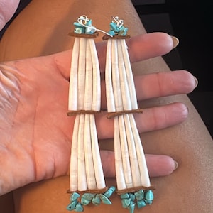 May include: A pair of long dangle earrings featuring vertical white cylindrical shell beads arranged in two tiers, separated and anchored by thin brown leather strips. The top and bottom sections are accented with small, irregular turquoise stones.