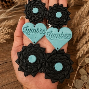 May include: Dangle earrings featuring a black textured star-shaped top piece with a light blue center, a light blue heart-shaped middle piece with the word "Jumbee" embossed in script, and a matching black star-shaped bottom piece with a blue center.