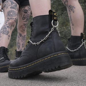 May include: Black leather platform boots with yellow stitching and a silver barbed wire chain. The boots have a lace-up design and a thick, ridged sole. The boots are worn by a person with tattoos on their legs.