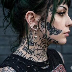 Könnte beinhalten: Eine Person mit mehreren Ohrpiercings und einem silbernen Zahn-Charm-Ohrring. Die Person hat ein Motten-Tattoo auf der Wange und Spinnennetz-Tattoos auf Gesicht und Hals. Die Person trägt ein schwarzes Oberteil.