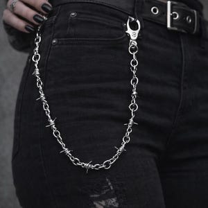 May include: A silver-coloured chain with barbed wire detailing hangs from black jeans. The chain is attached to the belt loop with a decorative clasp. The jeans have a distressed look with ripped areas.