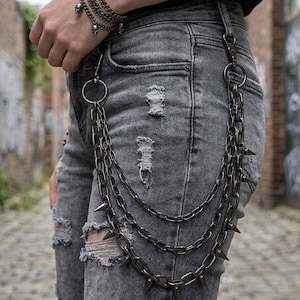 May include: A silver-toned, multi-strand chain with spike accents attached to a pair of distressed gray jeans. The chain is secured with a large ring and clasp. A matching skull bracelet is also visible.
