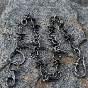 Op de afbeelding: Een zilverkleurige metalen ketting met een karabijnhaak en een decoratieve haak. De ketting is gemaakt van in elkaar grijpende metalen stukken, waarvan sommige op prikkeldraad lijken en andere op zeskantige moeren. De haak heeft een vogelontwerp.