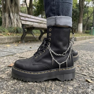 Może przedstawiać: Czarne skórzane buty bojowe z grubą platformą i metalowymi oczkami. Buty mają srebrne łańcuszki i kolce oraz są sznurowane. Noszone z podwiniętymi dżinsami.