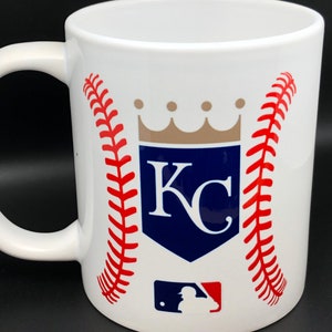 May include: White ceramic coffee mug with a baseball-themed design featuring the Kansas City Royals logo. The logo includes a blue shield with the letters "KC" in white, a gold crown above the shield, and red baseball stitching on either side. A smaller baseball logo with a red and blue silhouette of a baseball player is at the bottom.