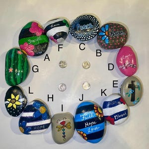 May include: A collection of painted rocks with inspirational words and floral designs. The rocks feature words like "Strength," "Peace," "Hope," and "Love." Some rocks have stripes, dots, or dragonfly designs. The rocks are arranged in a circular pattern.