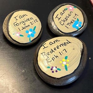 May include: Three smooth, rounded river stones painted with a tan circular center and black outlines. Each stone features hand-painted text: I am Forgiven 1 John 2:12, I am Chosen 1 Thes 1:4, and I am Redeemed Eph 1:7, accented with small colorful floral designs.