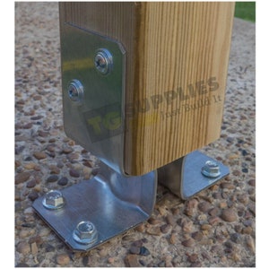 May include: Close-up of a metal bracket attached to a wooden post with screws. The bracket has a curved base and is secured to a concrete surface with screws.
