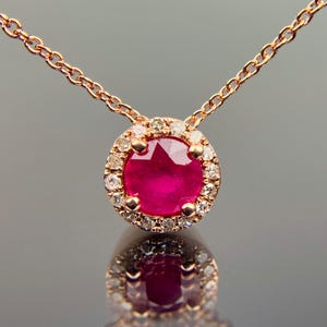 May include: A rose gold necklace featuring a round, vibrant red ruby gemstone pendant. The ruby is surrounded by a halo of small, sparkling diamonds. The pendant hangs from a delicate chain, creating an elegant and eye-catching piece of jewelry.