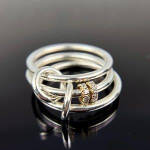 May include: A multi-band ring featuring three polished silver-toned bands interconnected by a single silver-toned loop. Two small, gold-toned circular spacers encrusted with tiny clear stones are threaded onto one of the silver bands.
