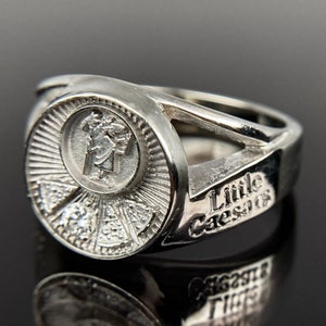 May include: A silver ring with a round design featuring a pizza slice and the words "Little Caesars" on the band.