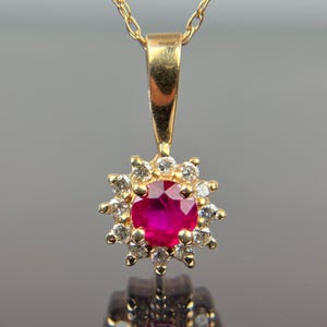 May include: A gold pendant necklace featuring a round, vibrant red gemstone surrounded by smaller, clear gemstones. The pendant is suspended from a delicate gold chain. The gemstones are set in a gold setting.