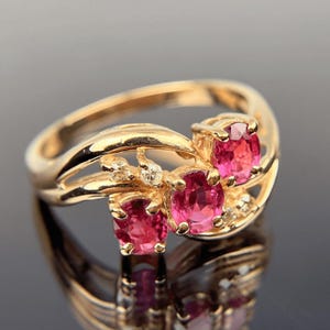 May include: A gold ring featuring three oval-cut, vibrant pink gemstones. The ring has a split shank design with small accent diamonds. The gemstones are set in a prong setting, and the ring is highly reflective.