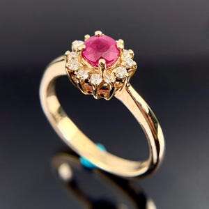 May include: A gold ring featuring a central, round, deep red ruby gemstone surrounded by a halo of small, clear diamonds. The ring's band is a polished gold color, and the setting is intricate.