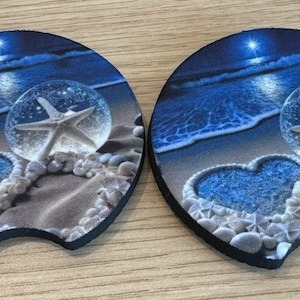 May include: Two round blue and white coasters with a heart shape and a starfish inside a glass orb on a sandy beach with waves in the background.
