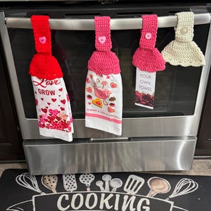 May include: Three crocheted kitchen towels hanging on a silver oven handle. The towels are pink, red, and beige. The towels have different designs and sayings. The pink towel says "Baked with Love". The red towel says "Love Grow". The beige towel says "Your Own Towel". The towels are hanging above a black and white kitchen mat that says "Cooking with Love".