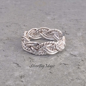 May include: A silver-colored ring with a braided design. The ring features interwoven strands, creating a textured and detailed appearance. The ring is displayed on a gray surface. The text "SilverBig Magic" is visible.
