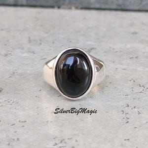 Könnte beinhalten: Ein silberner Ring mit einem großen, ovalen, schwarzen Edelstein. Der Ring hat ein schlichtes, poliertes Band und eine Lünettenfassung, die den dunklen Stein hält. Der Text "SilverBigMagic" ist sichtbar.