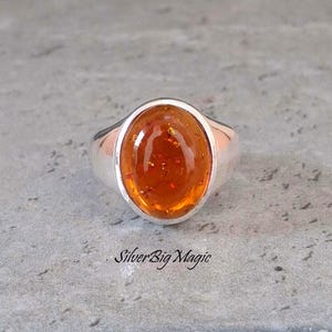 Peut inclure: Une bague en argent avec une pierre ovale de couleur ambre. La pierre a une teinte orange chaude avec des inclusions visibles. La bague est posée sur une surface grise. Le texte "Silver Big Magic" est visible.