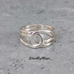 May include: A silver ring with a distinctive design. The ring has a central loop and multiple bands, creating an open, layered effect. The ring is set against a grey background, with the text "SilverBigMagic" visible.