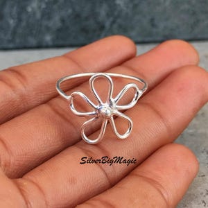 May include: A silver-colored ring with a flower design. The ring features six curved petals surrounding a central bead. The ring is held in a hand. The text "SilverBigMagic" is visible on the hand.