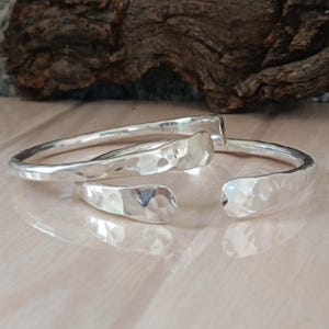 May include: Two silver cuff bracelets with a hammered texture. The bracelets feature a polished, reflective finish and an open design, with one bracelet slightly overlapping the other. The metal surface shows distinct indentations from the hammering.