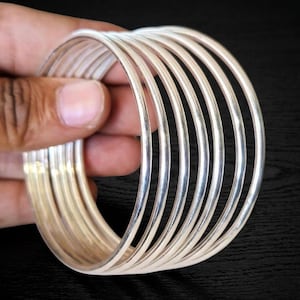 May include: A stack of silver bangles. The bangles are thin, round, and shiny, reflecting light. The bracelets are held in a hand against a dark background. The bangles are a simple, elegant accessory.