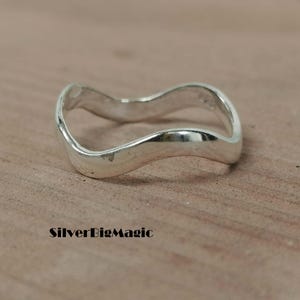 Peut inclure: Une bague en argent poli présentant un design ondulé continu et lisse qui se courbe de haut en bas. L'anneau possède une surface métallique réfléchissante. Le texte SilverBigMagic est imprimé en noir dans le coin inférieur gauche.