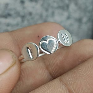 Op de afbeelding: Een zilveren ring met drie ronde bedels op een rij. De eerste bedel is voorzien van de letter I, de middelste bedel heeft een hartvorm en de derde bedel is voorzien van de letter U, wat samen I hart U vormt als een tekstuele boodschap.