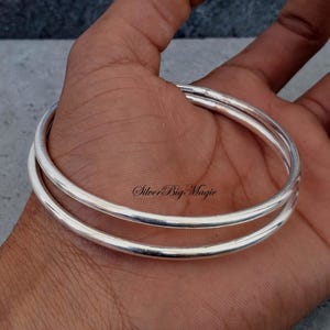 Puede incluir: Dos pulseras de plata, cada una con un diseño liso y redondeado. Las pulseras se sostienen en una mano, mostrando su superficie pulida y reflectante. El texto "Silver Big Magic" es visible.