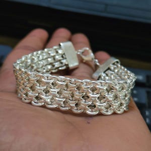 May include: A silver-toned bracelet featuring a multi-strand woven chain design with a polished metallic finish. The piece includes a rectangular metal end cap and a lobster claw clasp mechanism for secure fastening, showcasing a dense, interlocking link pattern.